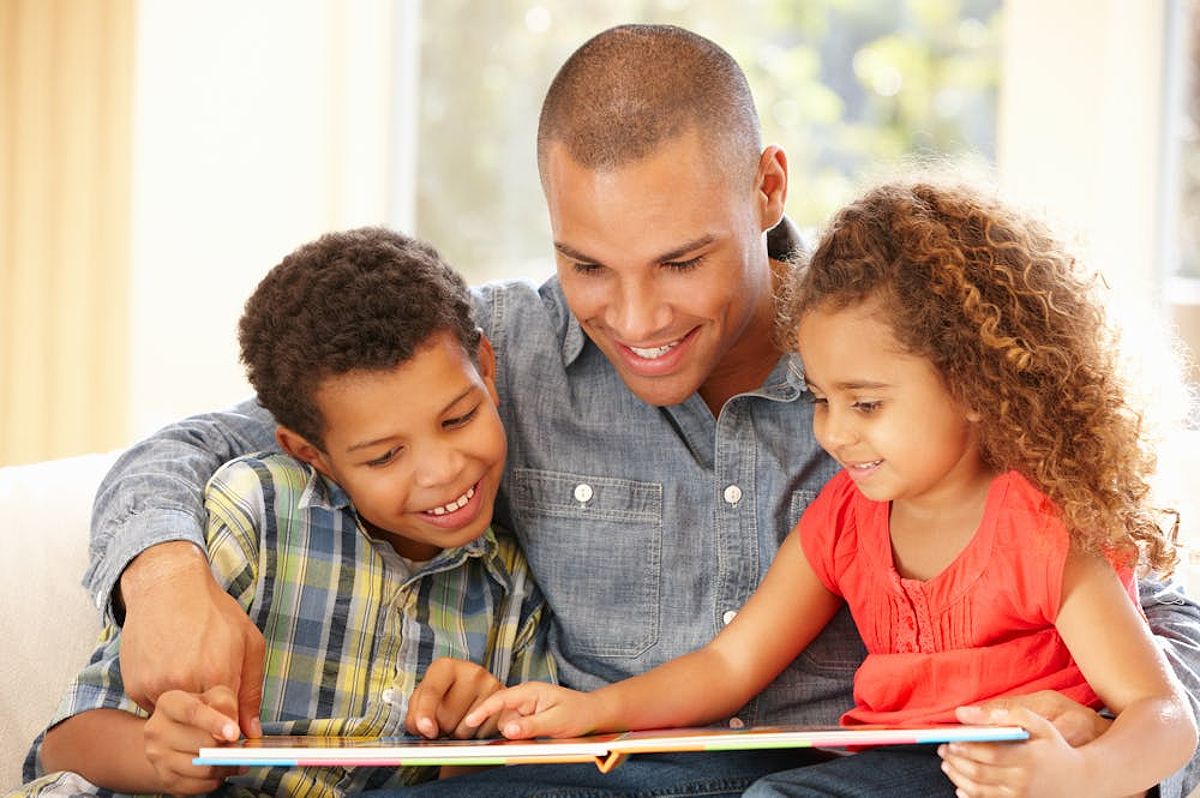 A man is reading a book to two children, smiling together indoors with a bright window in the background.